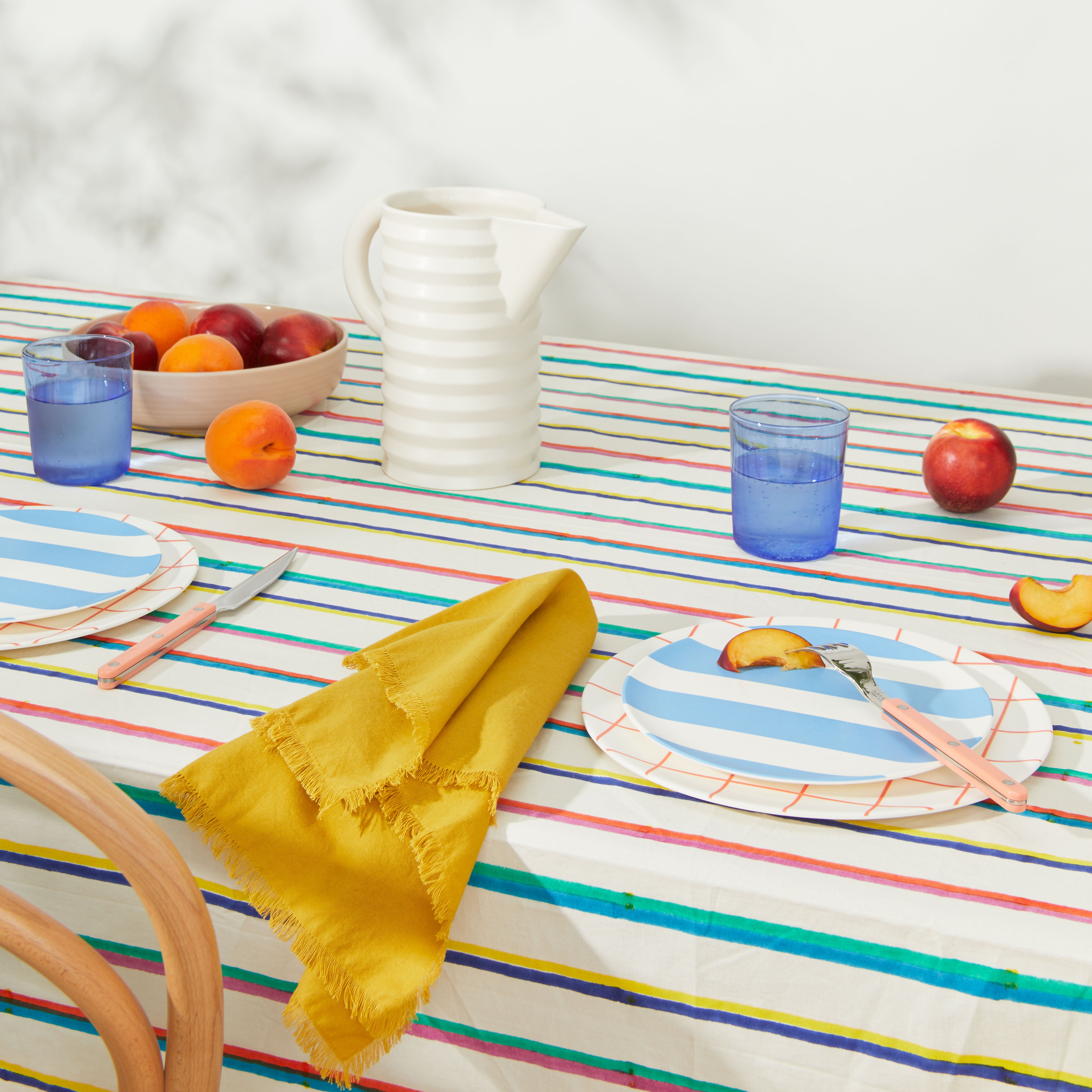 Picnic table setting with pitcher and blue glass tumblers on striped tablecloth.