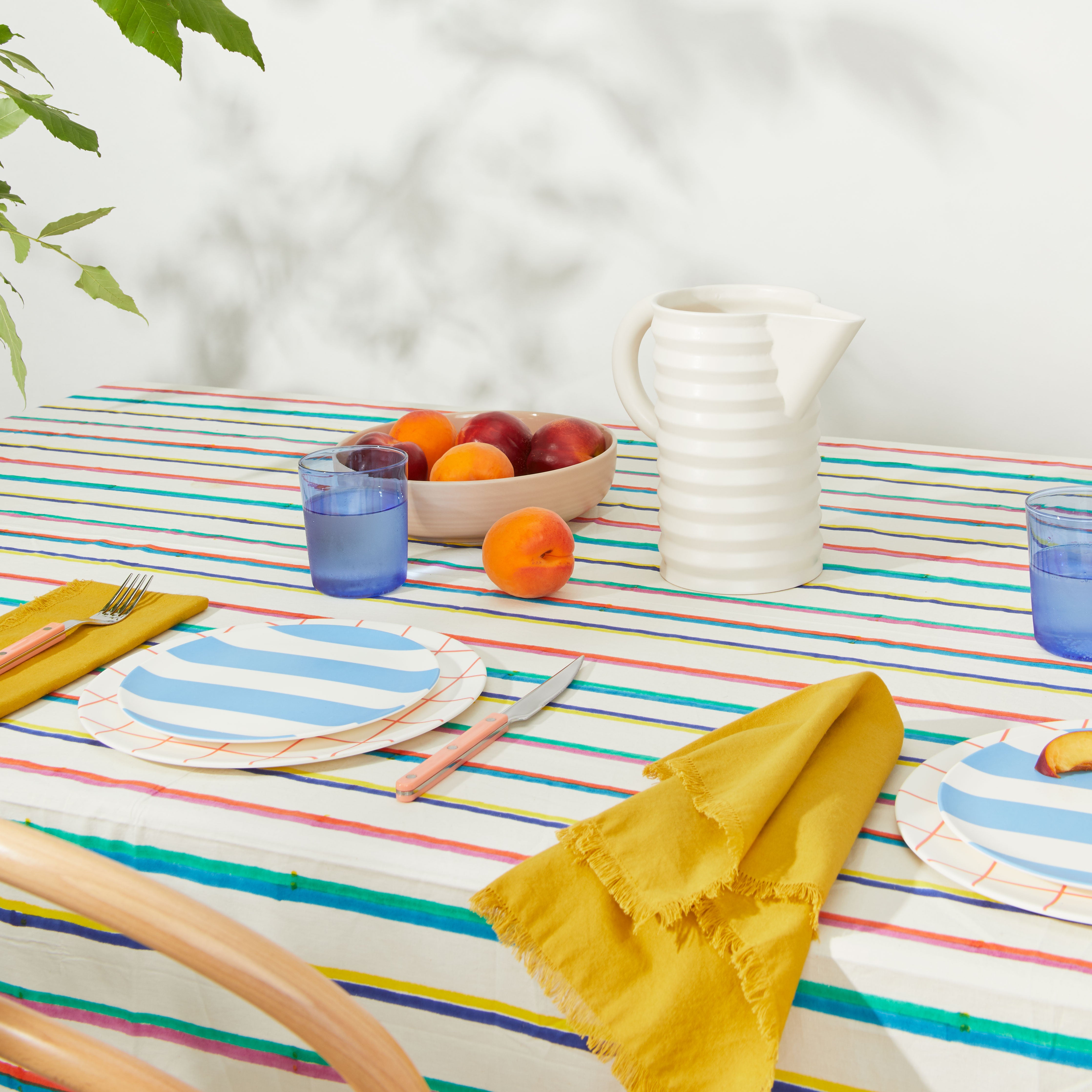 Picnic table setting with pitcher and blue glass tumblers on striped tablecloth.