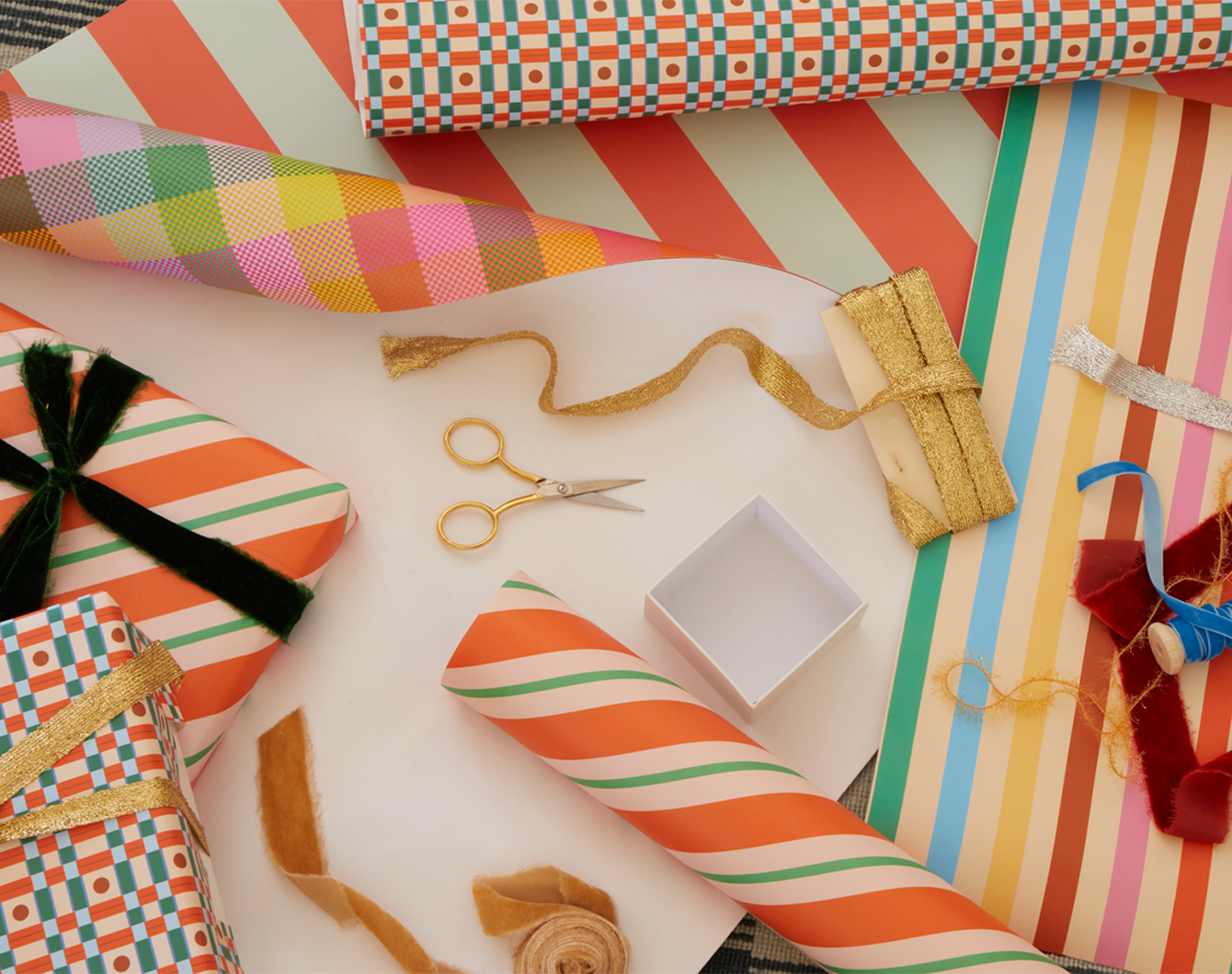 Various sheets of gift wrap with wrapped giftboxes, ribbon scissors, and ribbon on spools