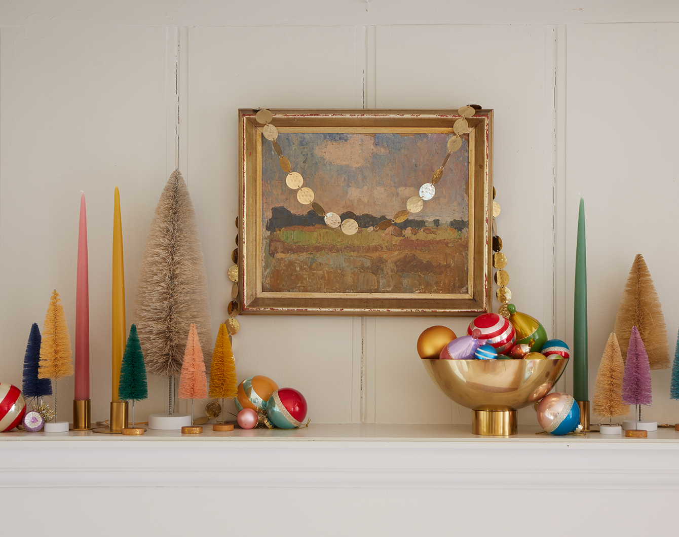 Mantel with framed artwork, candles, bottlebrush trees, various taper candles in brass candlestick holders, and brass footed bowl filled with ornaments.