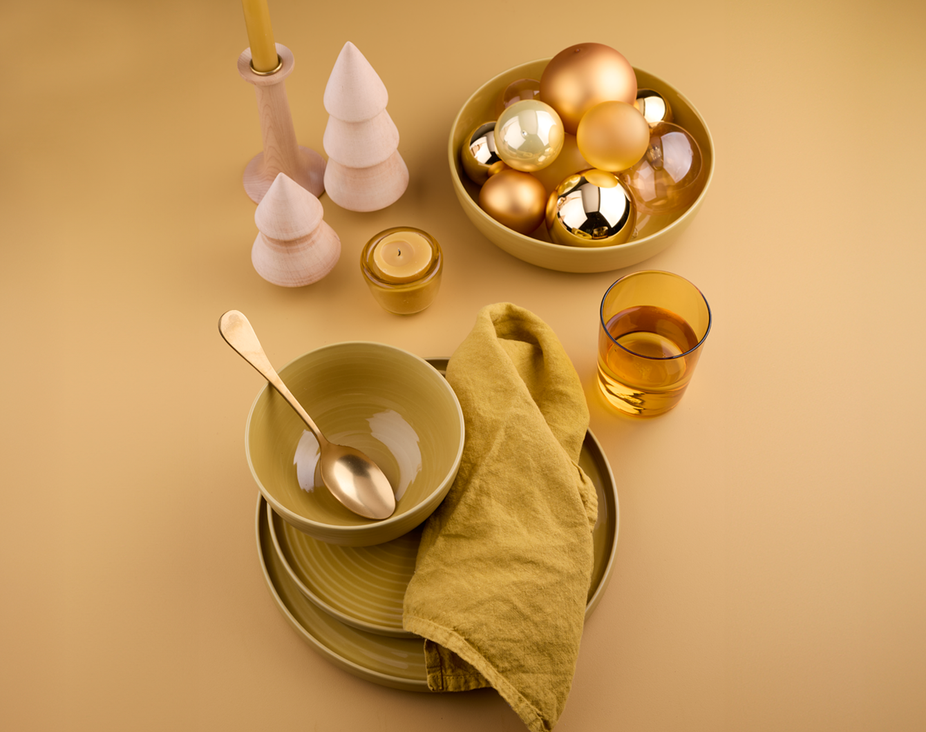 Yellow tabletop with mustard dinnerware, wooden trees, a single yellow glass, napkin, votive holder, and  taper candle holder