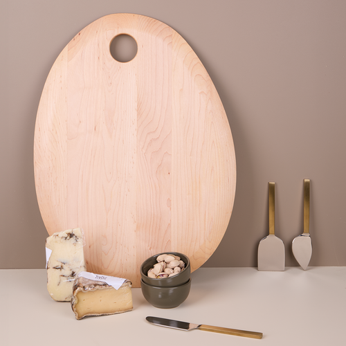 Maple pebble-shaped cutting board alongside an assortment of cheese, a set of three metal cheese knives, and a set of two Olive colored extra small bowls filled with nuts.