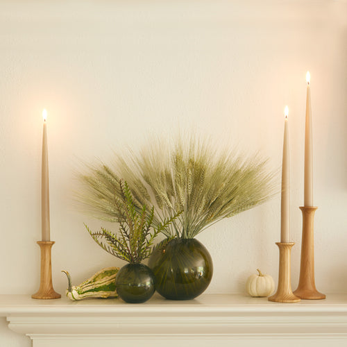 A mantel scene featuring a pair of Aurora Sphere Vases in Olive and a trio of stepped-height Simple Candle Holders in Oak with Taupe taper candles. Dried florals and seasonal gourds complement the pieces.