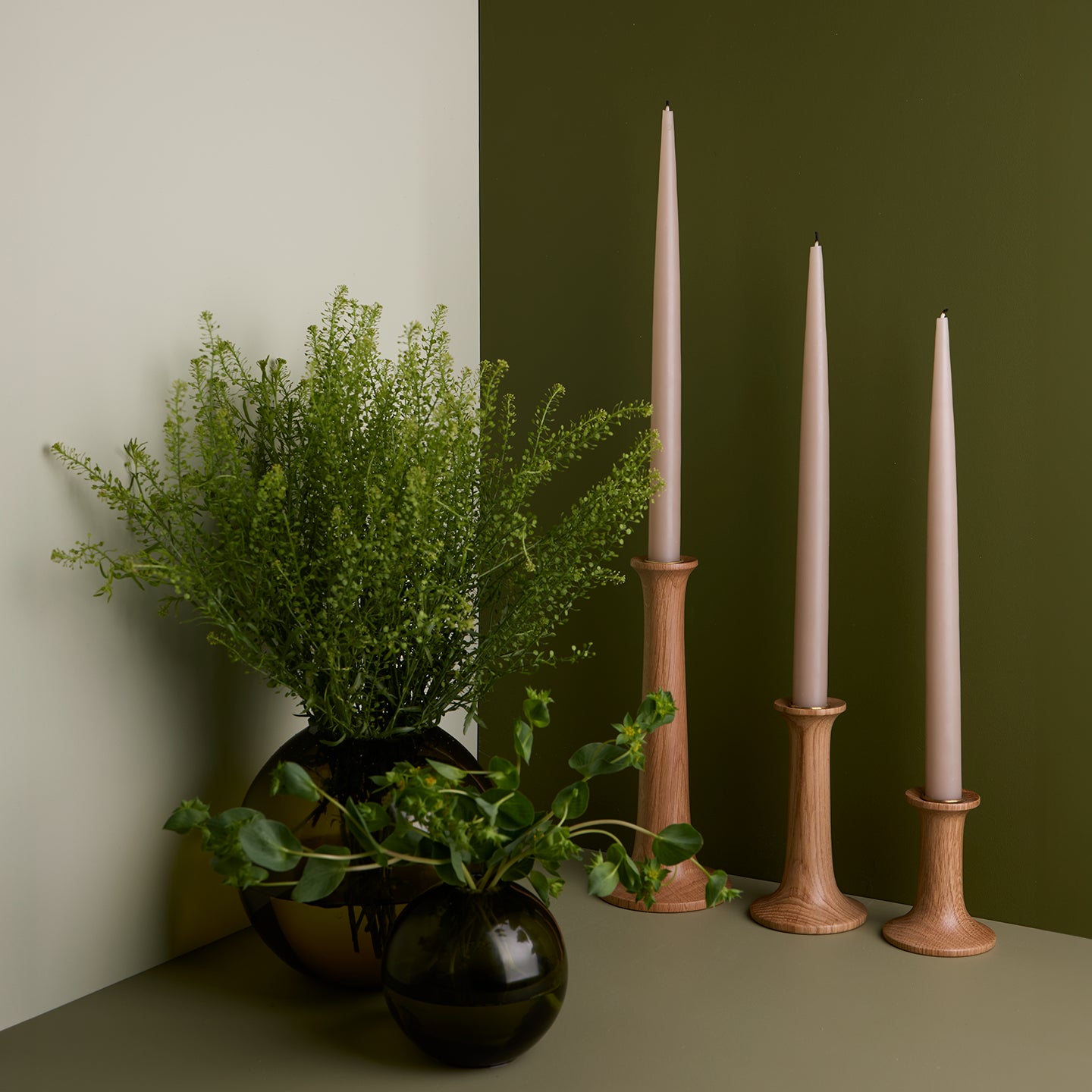 A pair of glass sphere vases filled with flowers and a trio of taper candle holders with taper candles. All in Olive tones.