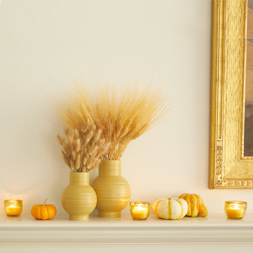 A mantel scene featuring a pair of Essential Ceramic Vases in Mustard and a trio of Aurora Votive holders in Amber with Honey votive candles. Dried florals and seasonal gourds complement the pieces.