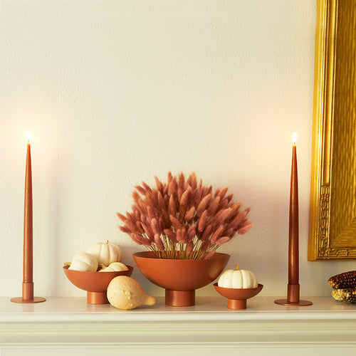 A mantel scene featuring a trio of Essential Footed Bowls in Terracotta and a pair of Essential Metal Candle Holders in Terracotta with Rust taper candles. Dried florals and seasonal gourds complement the pieces.