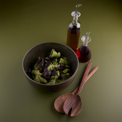 Essential Serving Bowl in Olive filled with leafy greens, alongside walnut wood serving set and glass oil and vinegar bottles.