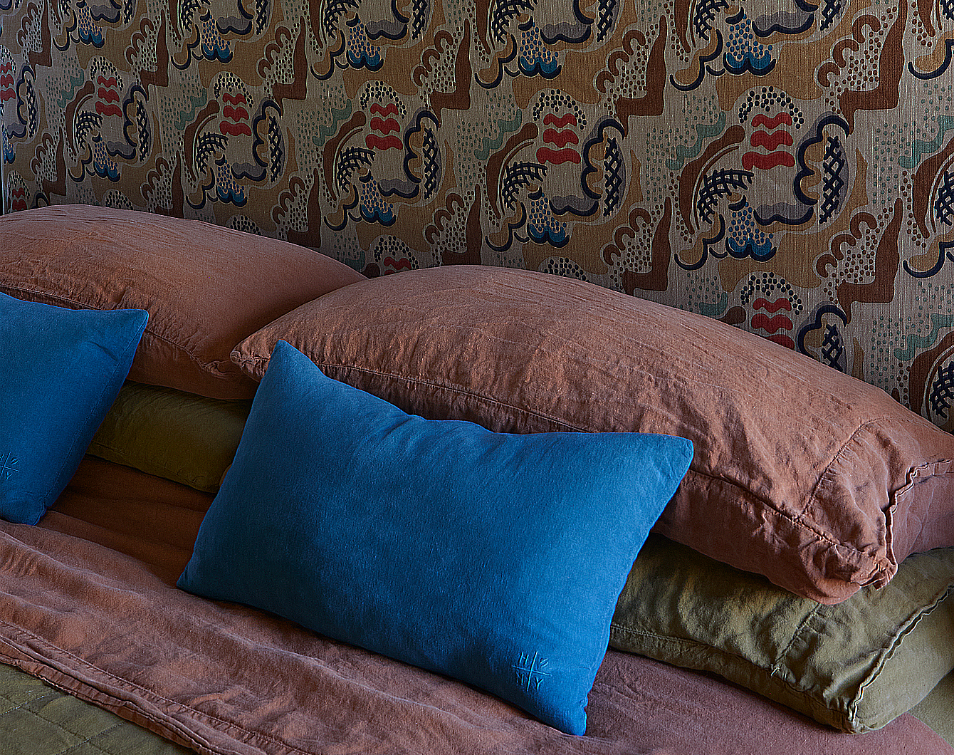 Bed with linen bedding and stacks of pillows against a patterned upholstered headboard.