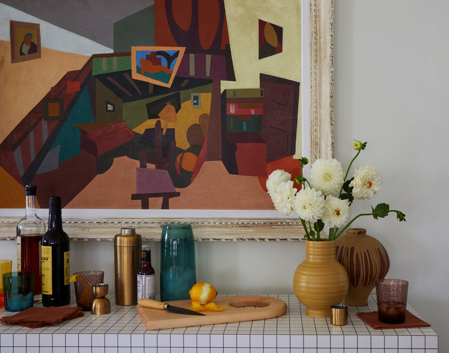 Home bar on console table with glassware, cutting board, vase of flowers, cocktail napkins, and glassware, with a colorful framed painting hanging above.