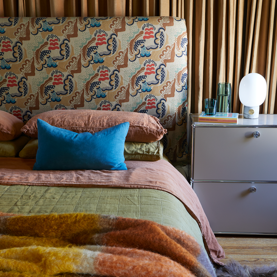 A Bedroom with a patterned headboard, linen pillows in various colors, a sage quilt, and a mohair throw sits next to a grey bedside table with a lamp and glass with a pitcher