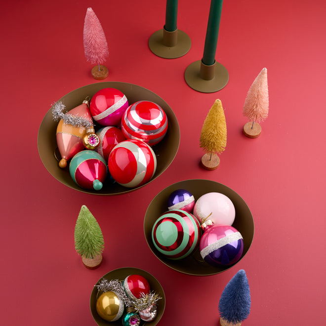 A trio of footed bowls filled with colorful holiday ornaments. Nearby a pair of candle holders with green taper candles. Arranged with a group of bottlebrush trees.