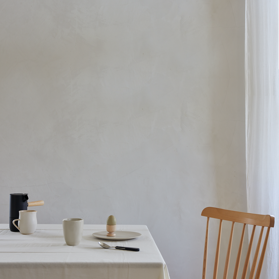 A minimalistic dining setup with a white tablecloth, a wooden chair, a black coffee pot, a white mug, a plate with an egg in an egg cup, and a knife and spoon on the table.