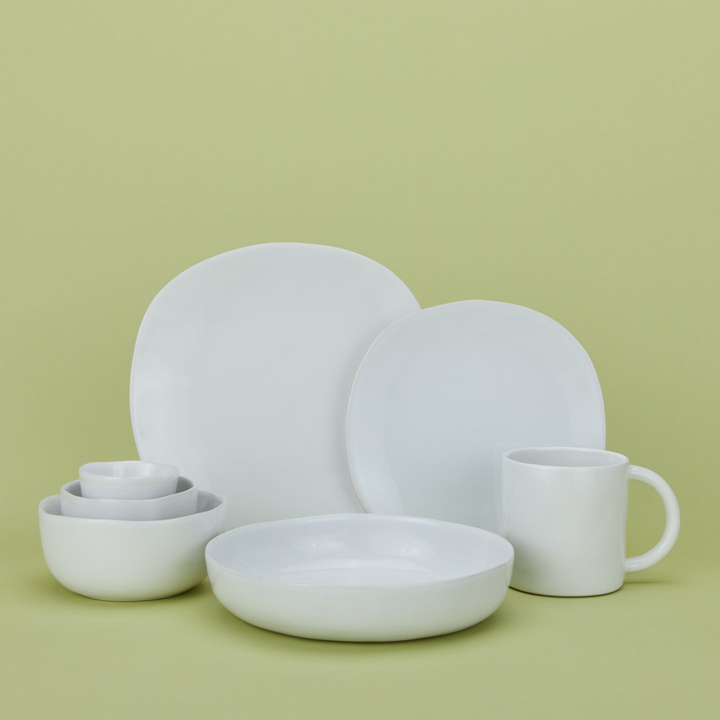 Group of white Strata dinnerware pieces including mug, various sized plates and various sized bowls against a green background.