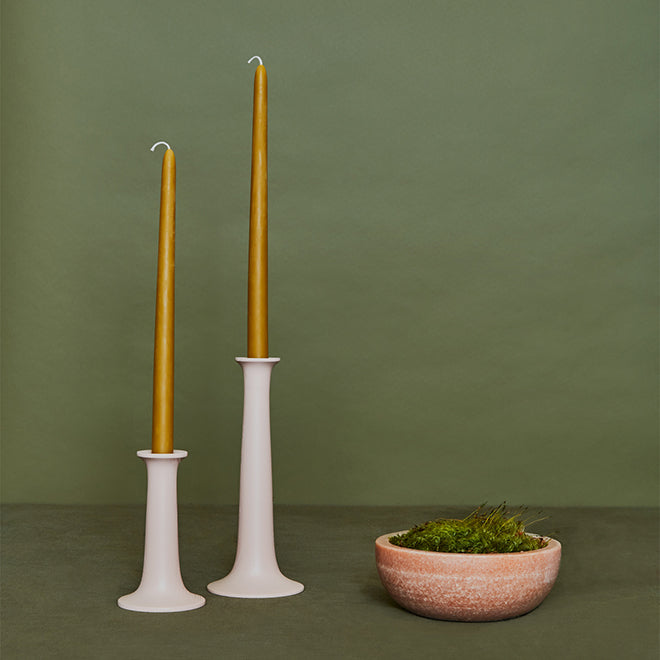 Two lit mustard-colored taper candles in white holders beside a beige bowl filled with moss, all set against a green background.