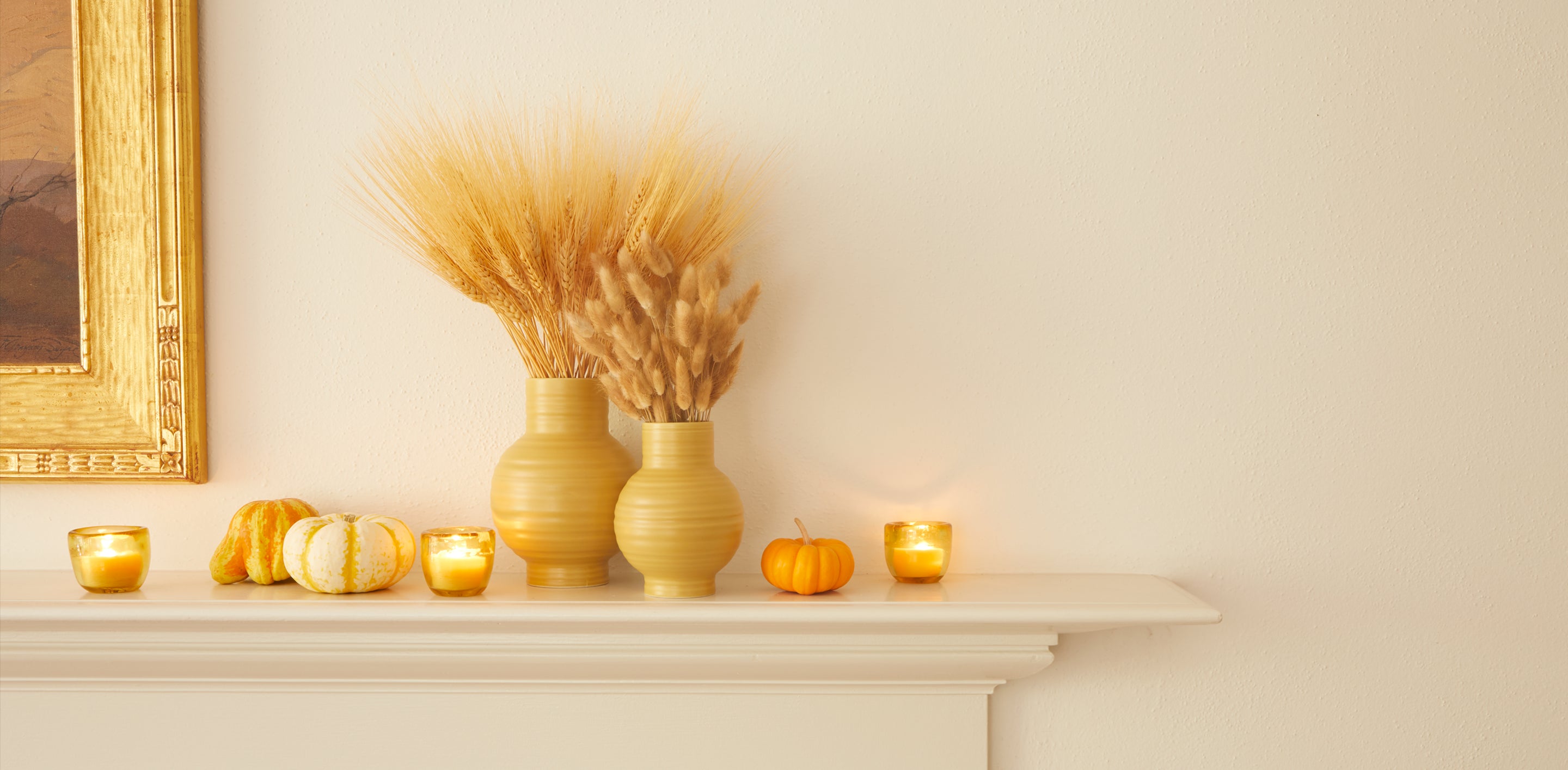 A bright mantel with vases of wheat, flanked by lit votives in glass holders, and mini pumpkins and gourds