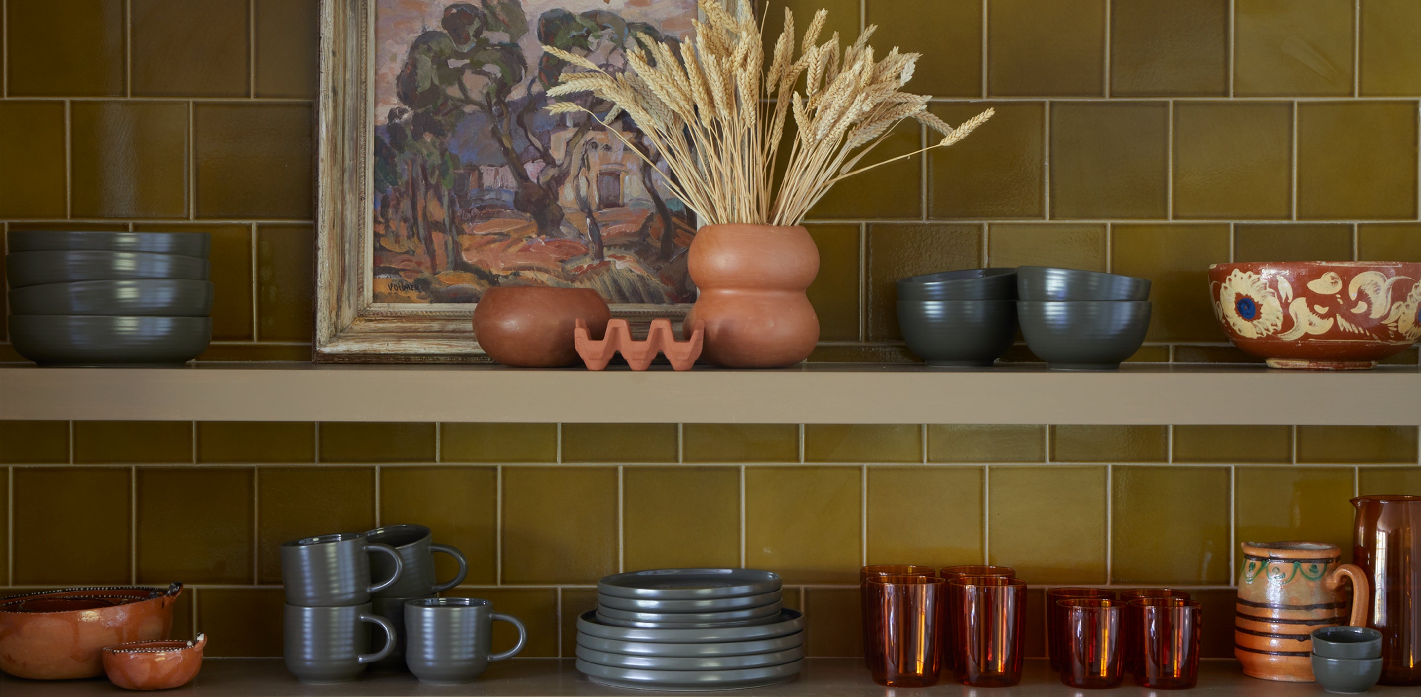 A tiled kitchen wall with stacks of dinnerware, glass ware and pottery with artwork placed in the middle of a shelf