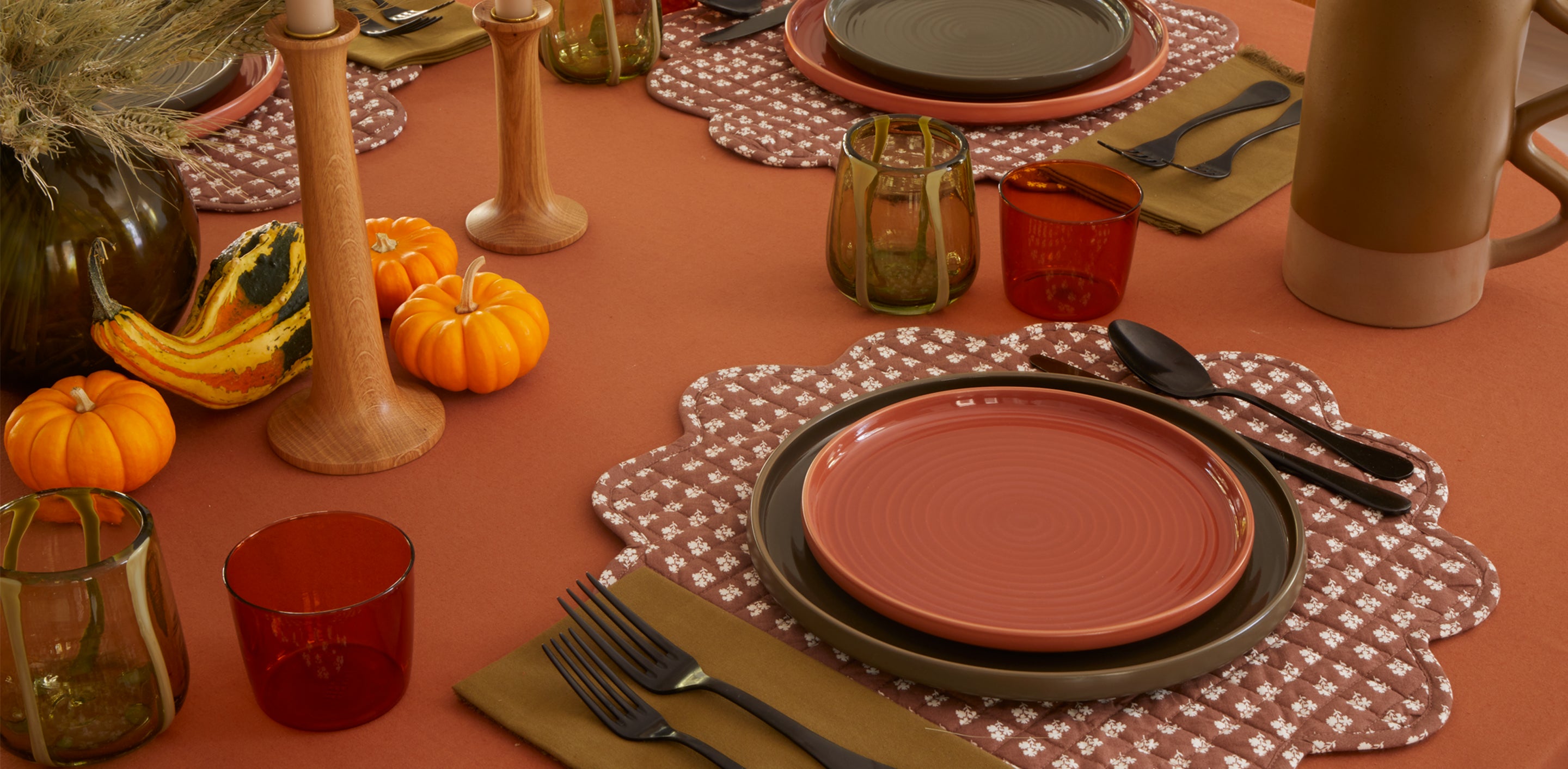 A thanksgiving place setting with quilted placemats, plates, flatware, glassware, and a pitcher, pumpkins and gourds are placed around a vas containing dried florals