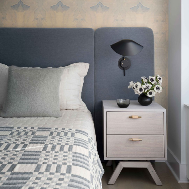 A tidy bedroom with a blue upholstered headboard against a wall covered in light blue and ivory wallpaper, featuring a blue plaid blanket, a grey throw pillow, a bedside table with a vase of flowers, and a black wall-mounted lamp. 