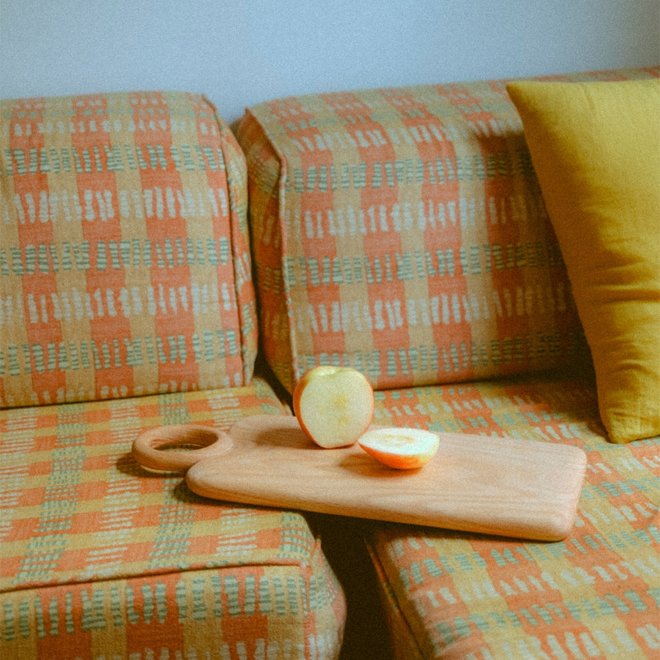 A cutting board with two halves of an apple and a yellow throw pillow sit atop a sofa upholstered in an irregular striped fabric in gold, orange and green tones.