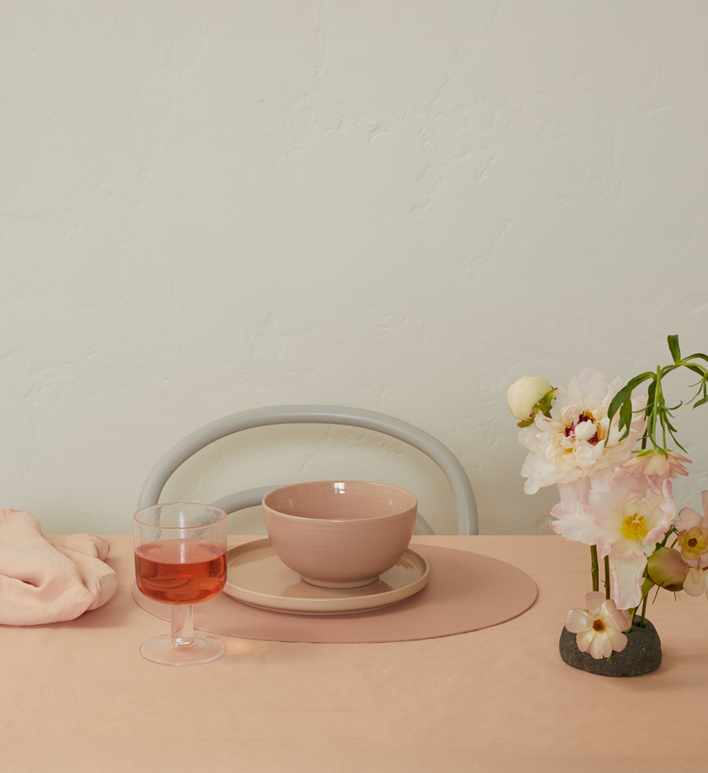 A tablesetting featuring a placemat, salad plate, bowl, and linen napkin, all in blush tones. A stemmed glass is full of rose. Nearby sits an ikebana style flower arrangement in a river rock floral frog. 