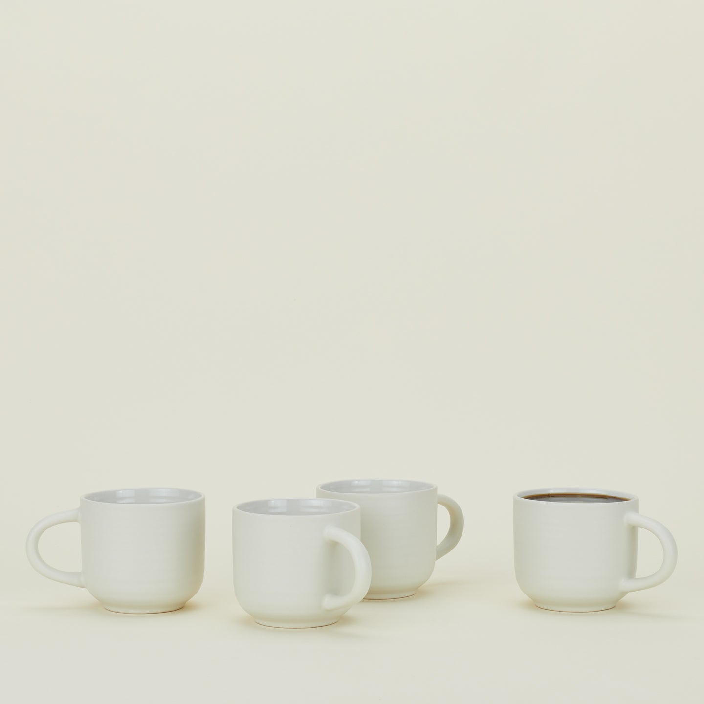 Four white ceramic mugs are arranged in a row on a plain light background. The mug on the far right is filled with a dark liquid.