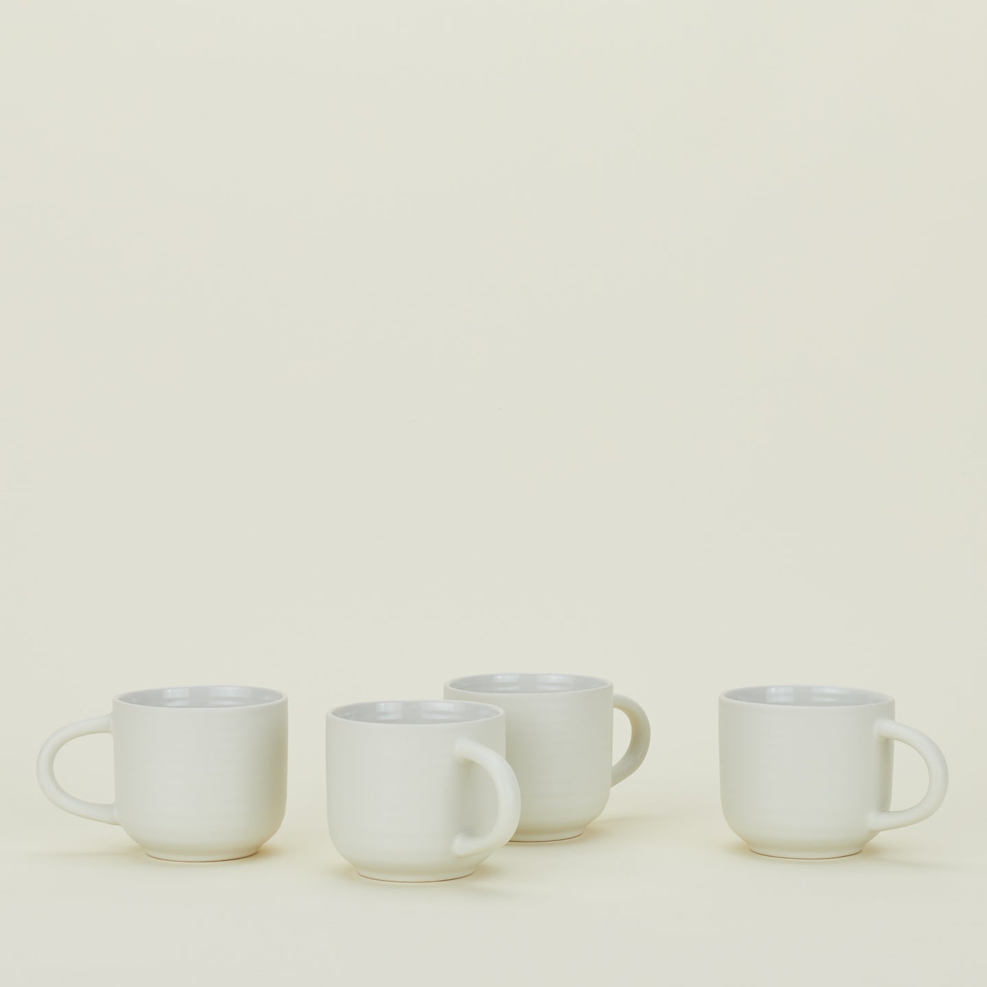 Four white ceramic mugs with handles are arranged in a row against a plain, light background.