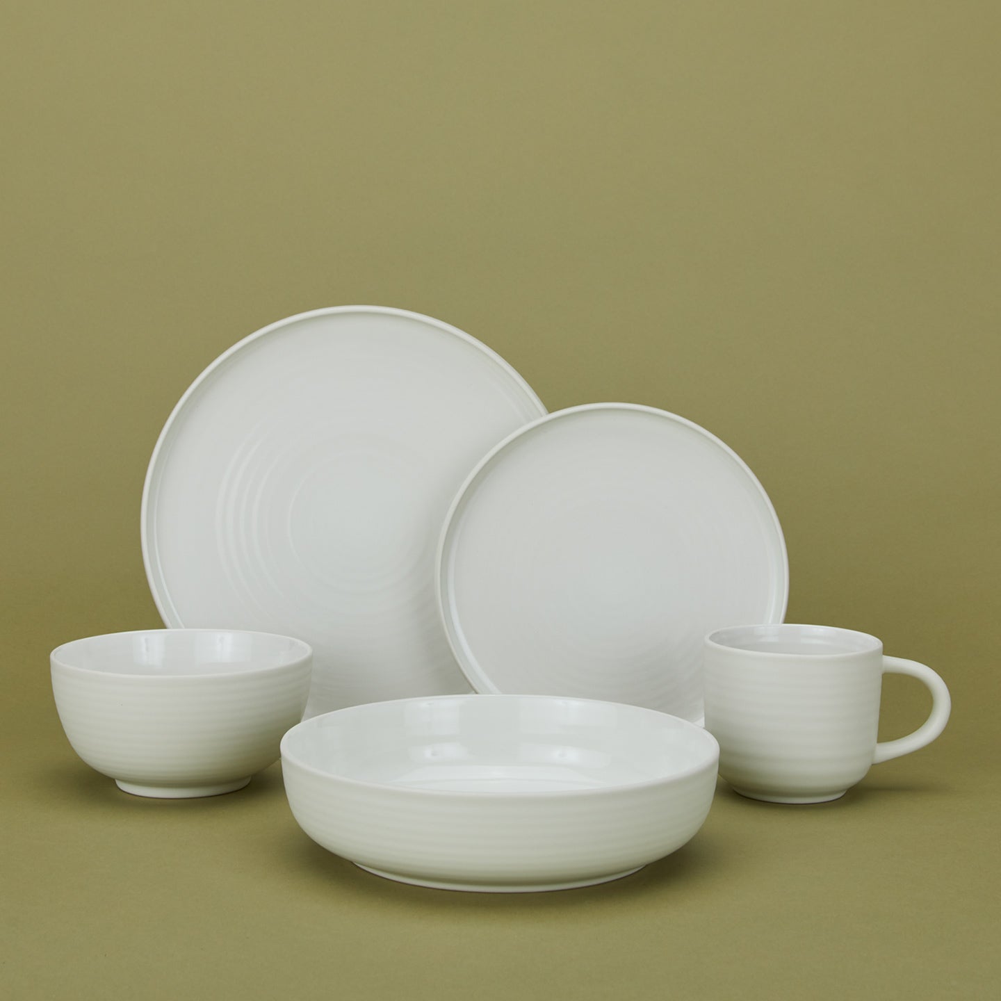 A set of white ceramic dishware including two bowls, two plates, and a mug, arranged in front of a green background.