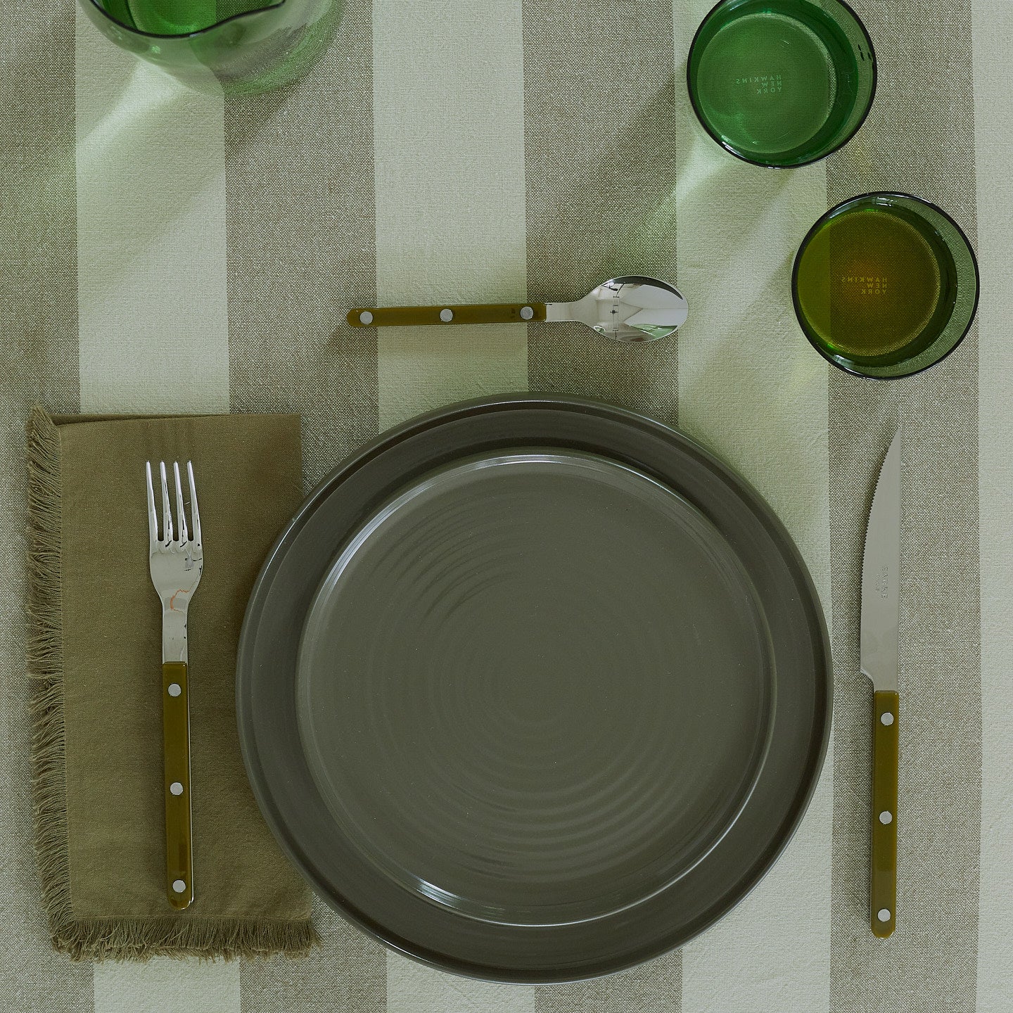 Placesetting of olive green Essential dinnerware on green striped tablecloth.