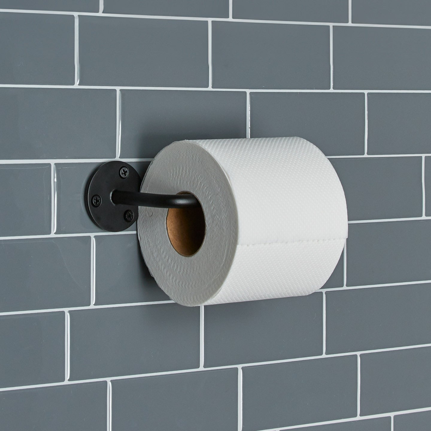 Essential toilet paper holder in Black with a roll of toilet paper on tiled wall.