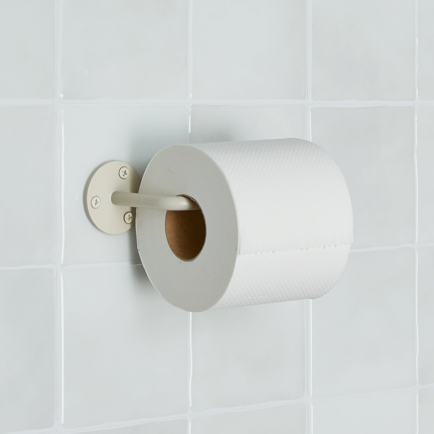 Essential toilet paper holder in Ivory with a roll of toilet paper on tiled wall.