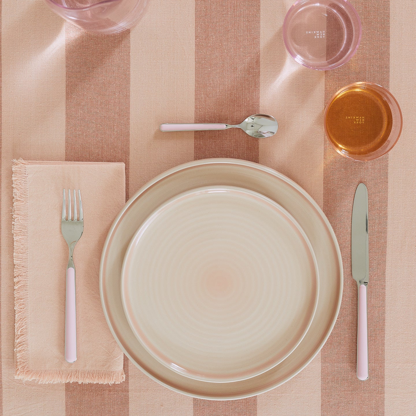 Placesetting with Essential Dinner Napkin in Blush on pink striped tablecloth.