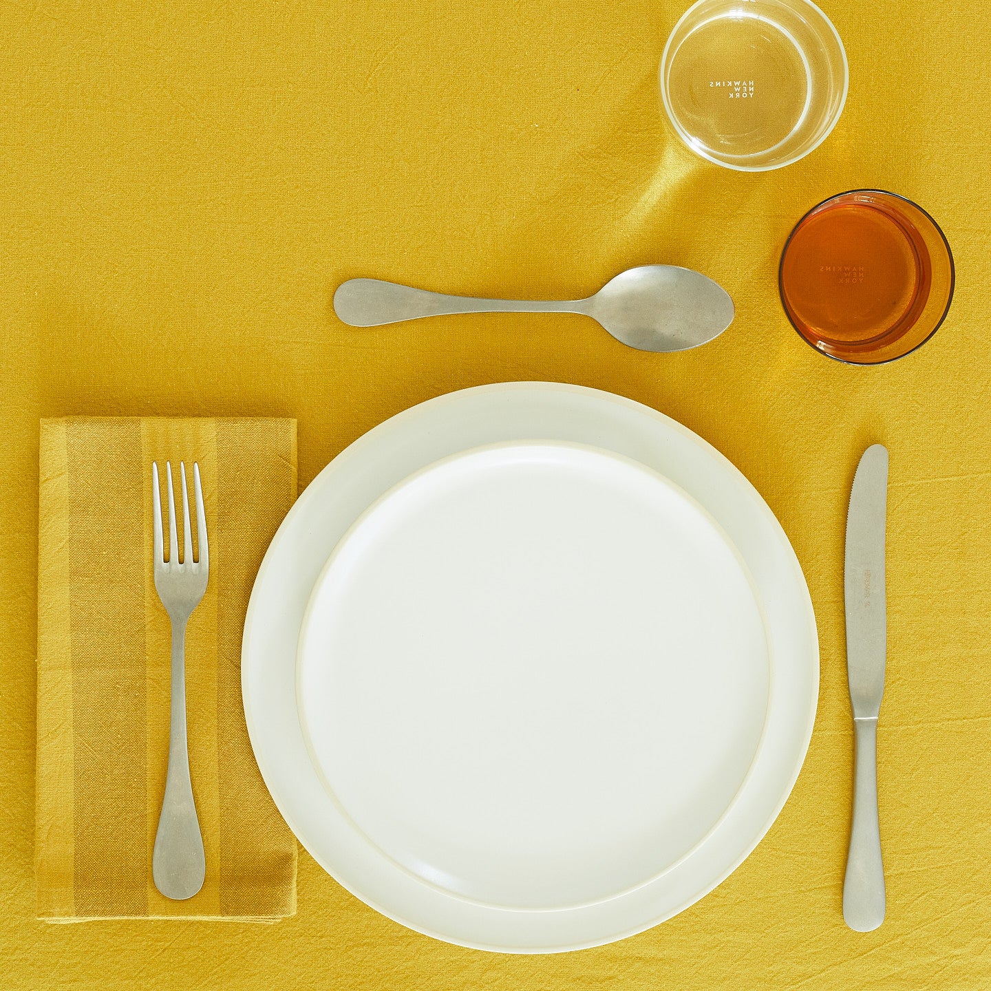 Placesetting with Essential Striped Dinner Napkin in Mustard and Bronze on yellow tablecloth.