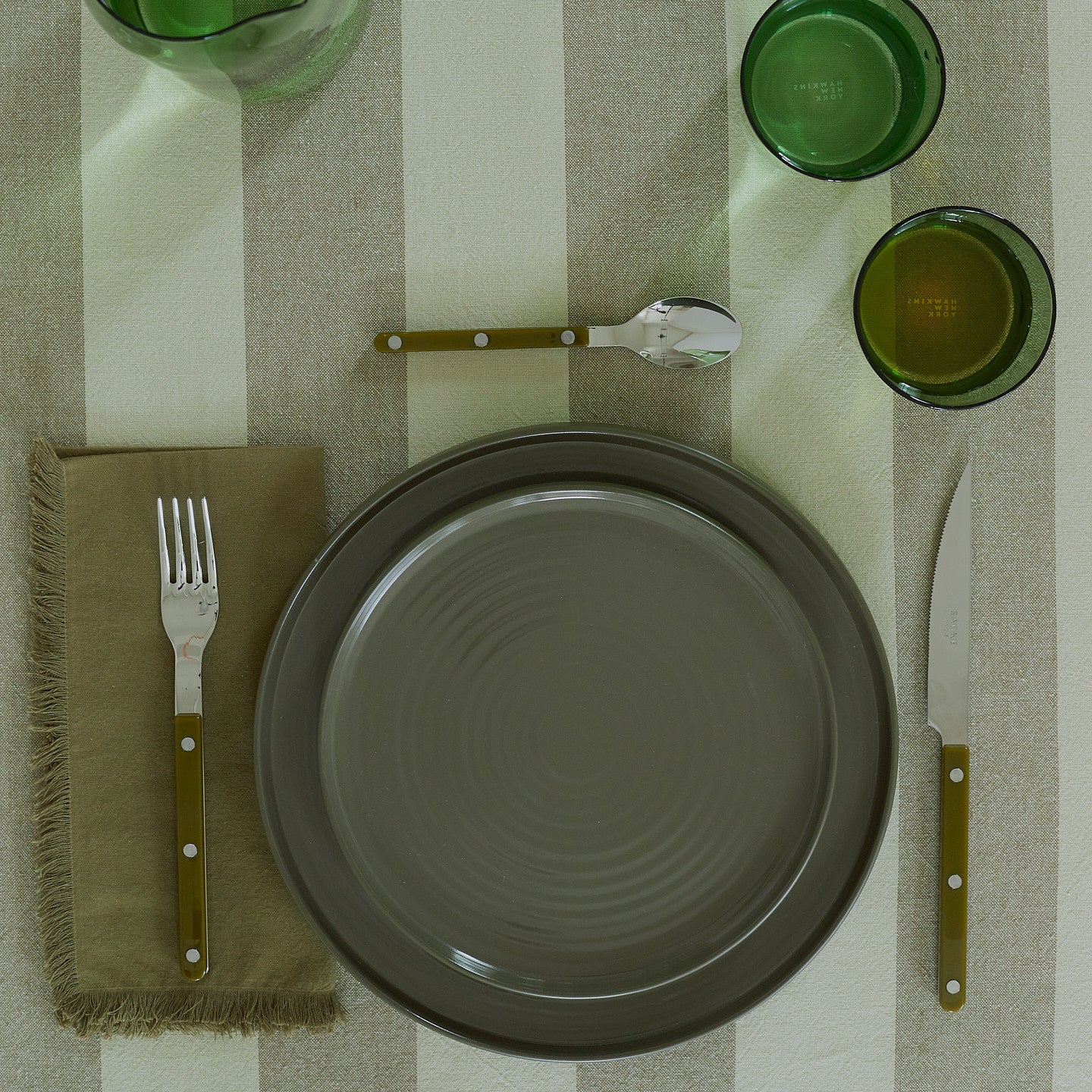 Placesetting with Essential Dinner Napkin in Olive on green striped tablecloth.