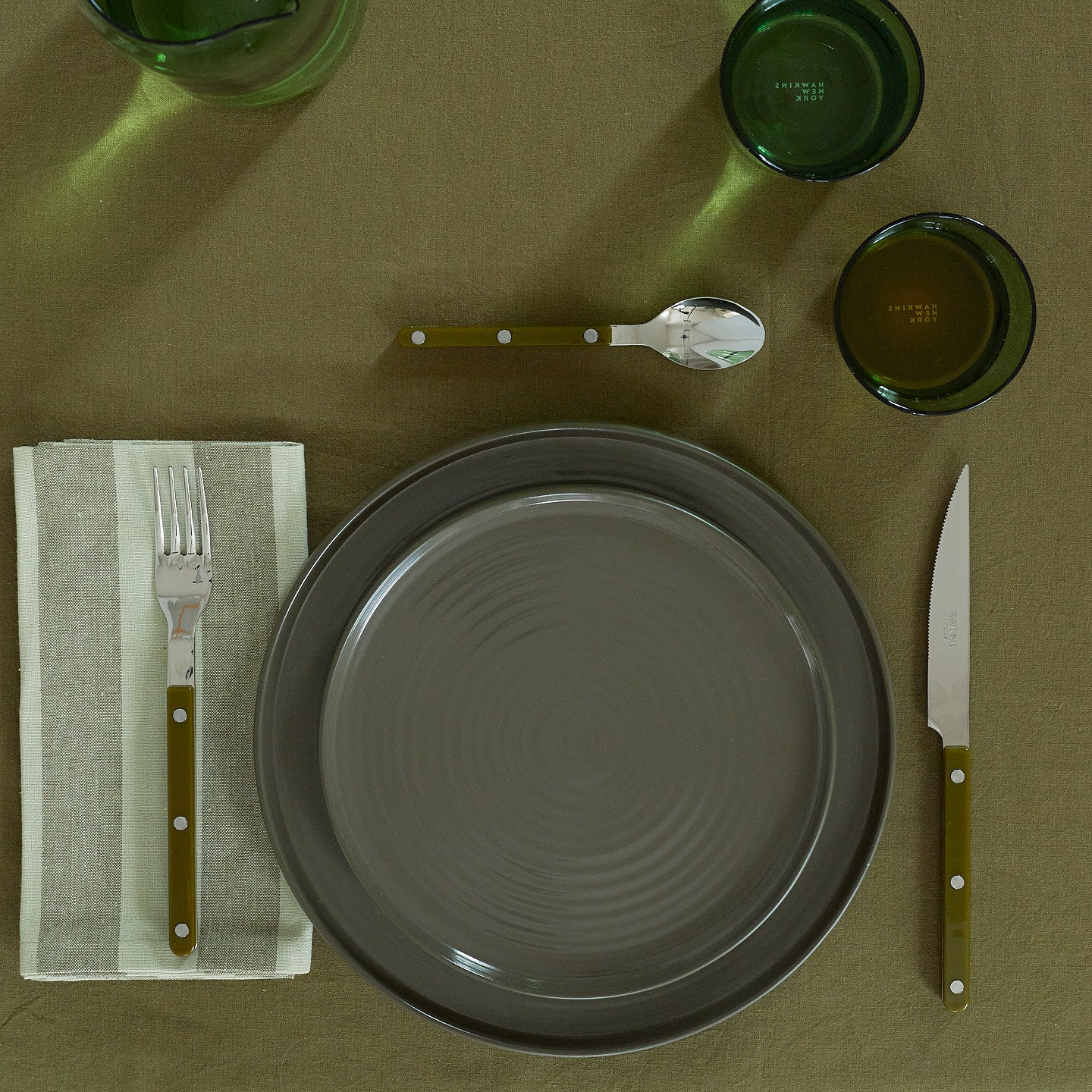 Placesetting with Essential Striped Dinner Napkin in Olive and Sage on green tablecloth.