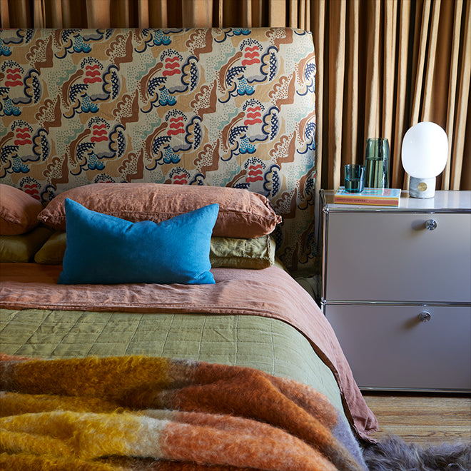 Bedroom scene with layers of linen bedding against a patterned upholstered headboard. A plaid mohair throw rests at the foot of the bed. A book, drinking glass, and carafe sit next to a bedside lamp on the nightstand.