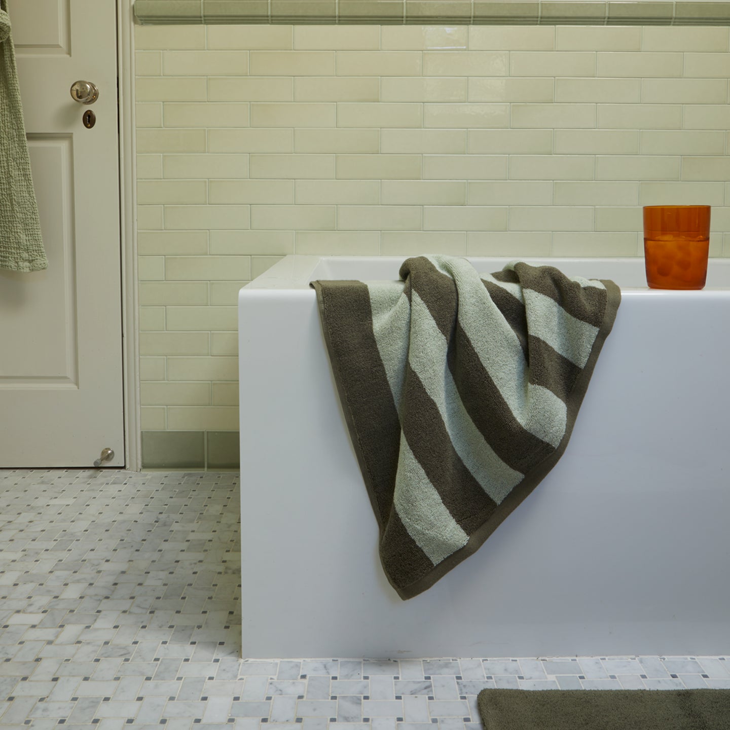 A striped towel draped over the side of a bathtub with a drinking glass resting on the edge.