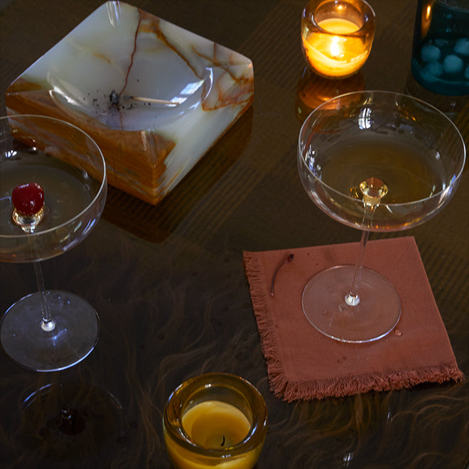 An assortment of votive candles, nearly empty cocktail glasses, and a marble ashtray with a burned match arranged on a glass coffee table sitting atop a sheepskin rug.