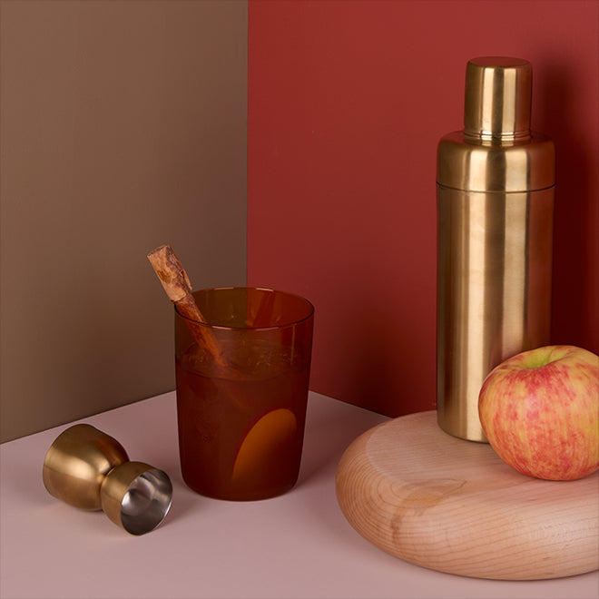 Cocktail garnished with cinnamon stick and apple slice next to brass finish jigger and shaker atop a wood chopping block.