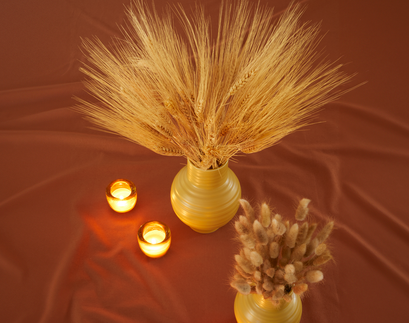 A pair of mustard colored ceramic vases filled with dried florals arranged with a grouping of amber glass votive holders, all resting on a terracotta colored tablecloth.