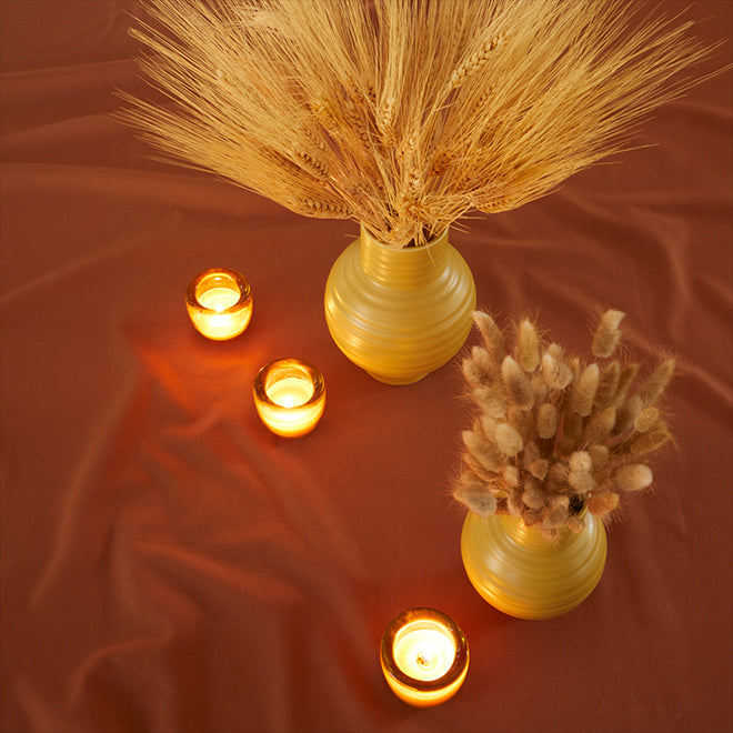 A pair of mustard colored ceramic vases filled with dried florals arranged with a grouping of amber glass votive holders, all resting on a terracotta colored tablecloth.