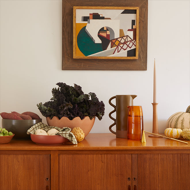 Various serving bowls filled with produce, a pair of pitchers, taper candle, douter, and seasonal gourds arranged atop a wood sideboard.