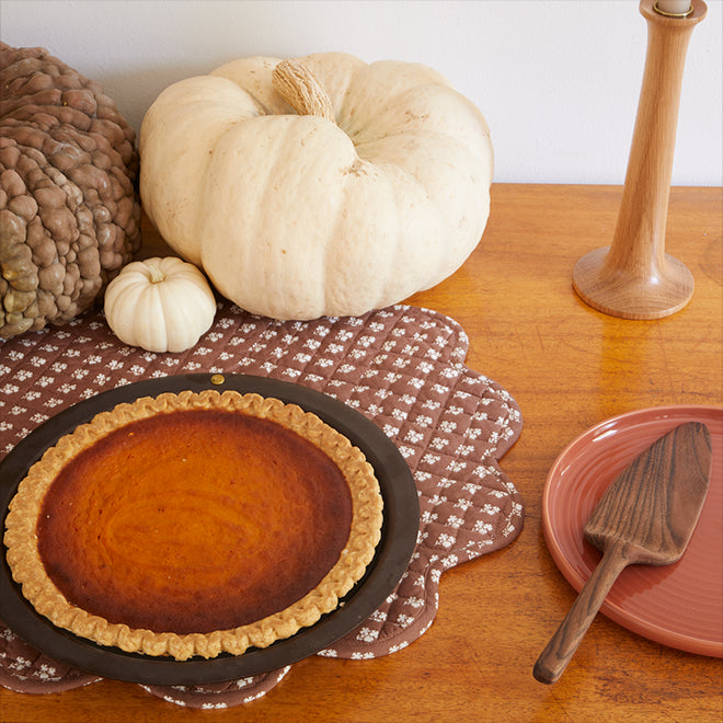 A pumpkin pie in a spun iron pie dish arranged alongside a group of pumpkins and gourds. Nearby a side plate and walnut pie server.