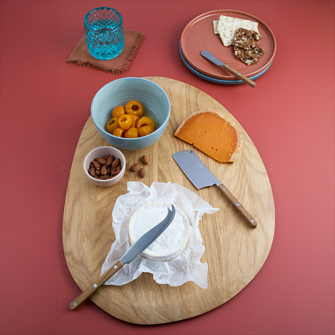 Wooden cutting board arranged with a collection of cheese knives along with various cheeses, crackers and nuts. Nearby a stack of side plates and a cocktail glass on a cotton coaster.