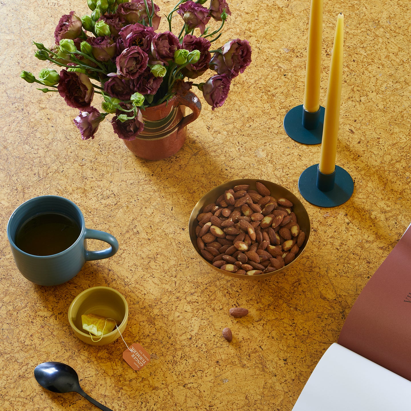 Mug with tea bag, spoon, bowl of nuts, vase of flowers, a pair of taper candles, and a book arranged on a cork coffee table.