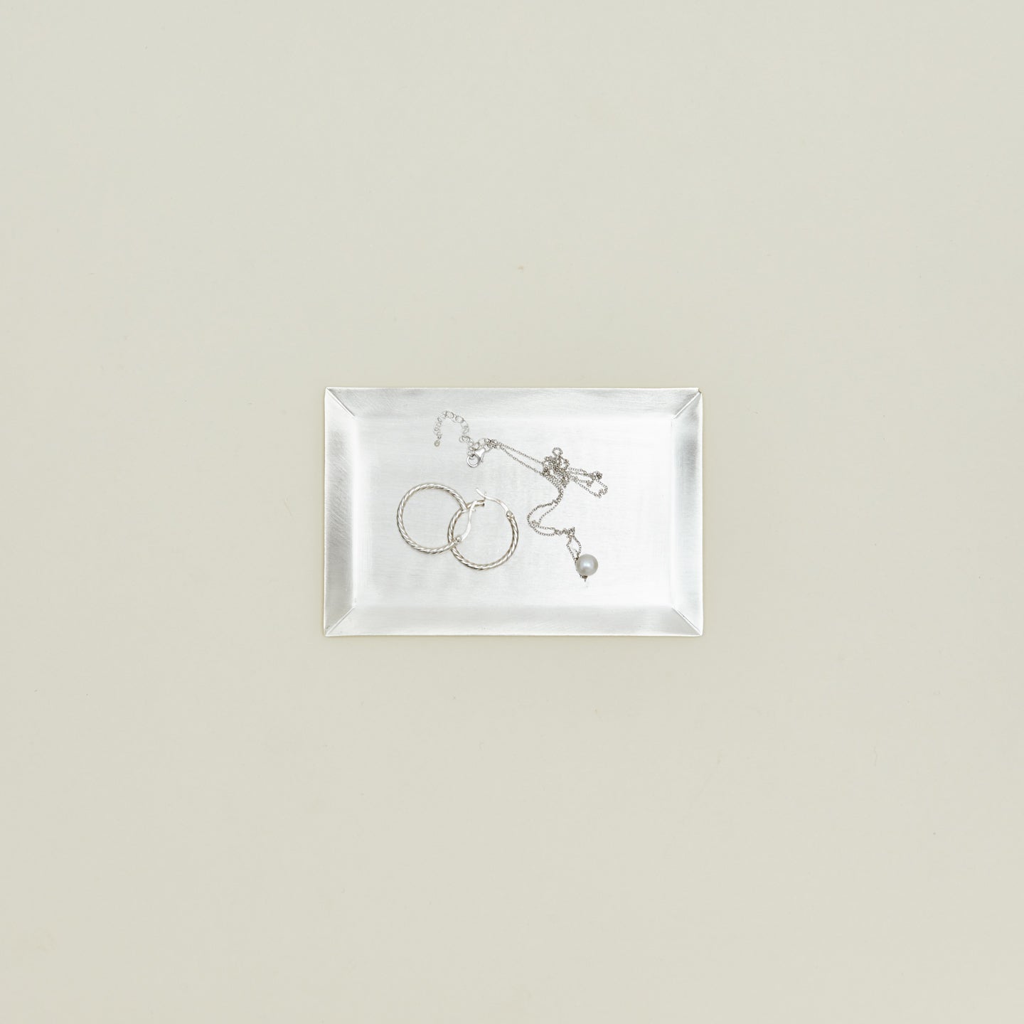 Overhead view of rectangular silver tray with a pair of hoop earrings and a pearl necklace inside.