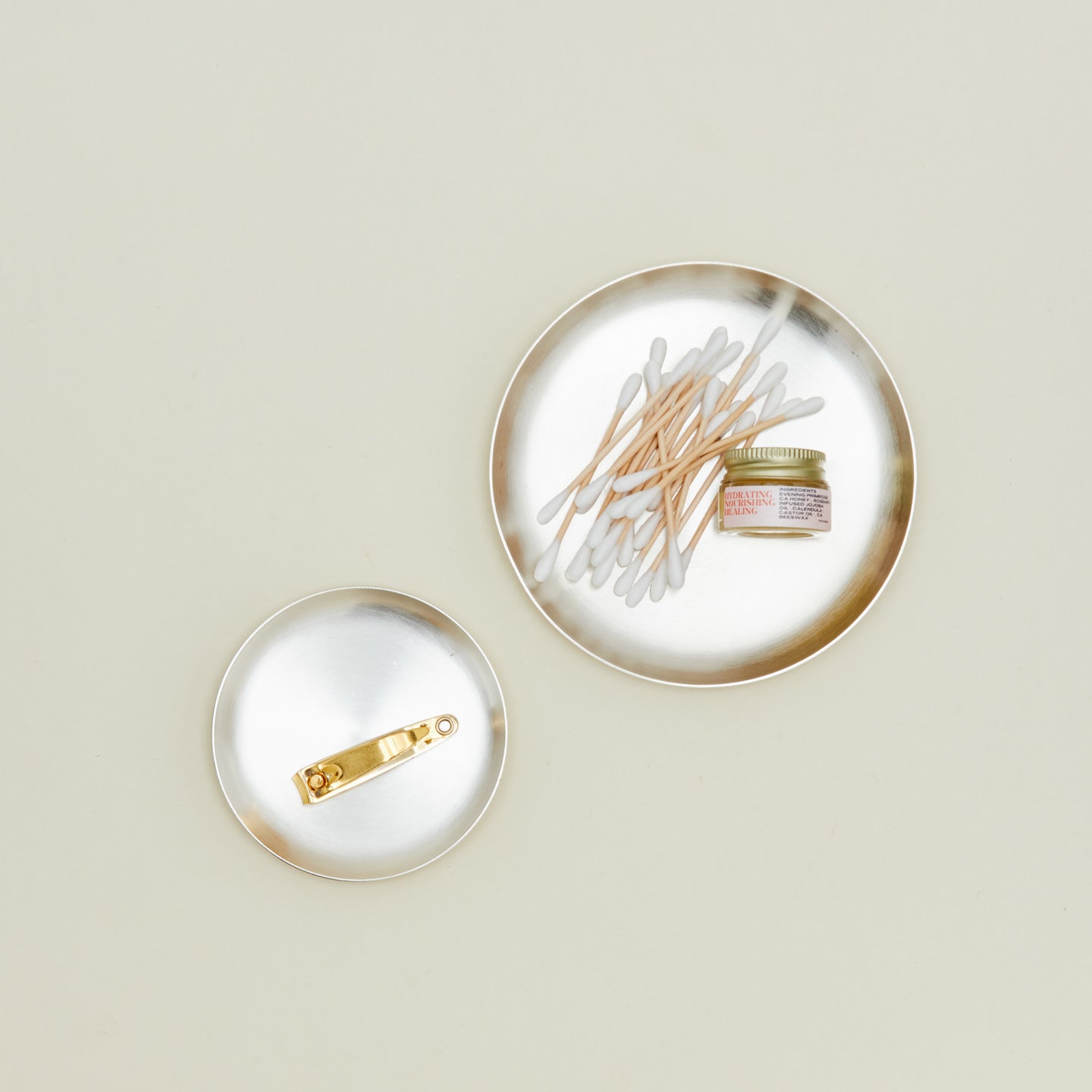 Overhead view of small and medium round silver plate with nail clippers in the small, and cotton swabs and lip balm in the large