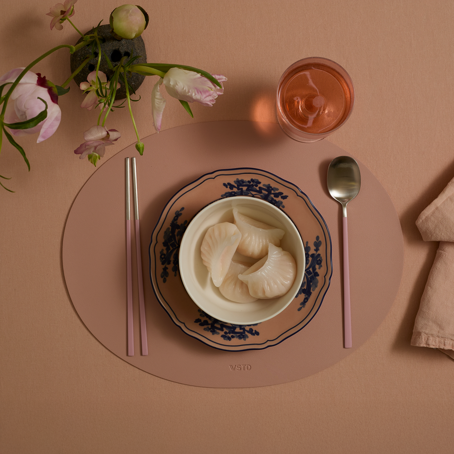 A small bowl of dumplings on top of a Ginori 1735 plate arranged on a blush oval-shaped placemat. On either side a pair of chopsticks and spoon with blush handles. Nearby a blush linen napkin, blush drinking glass, and an ikebana-style flower arrangement.