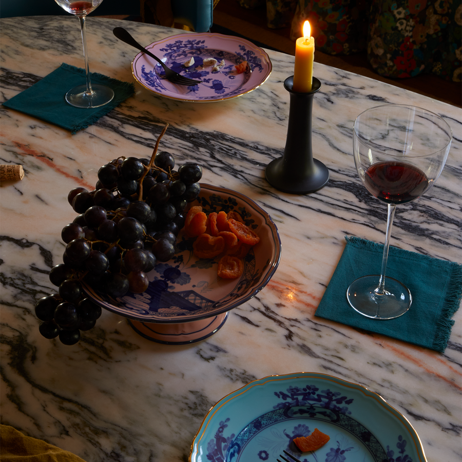 Ginori plates and fruit stand arranged on a marble tabletop with cloth napkins, fruit, wine glasses, and a lit taper candle.