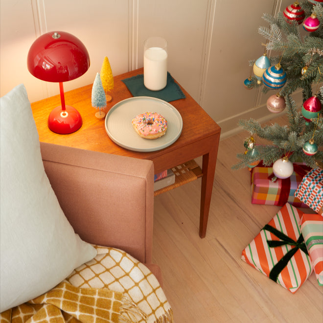 An armchair draped in a throw sits near a decorated Christmas tree with wrapped presents underneath. Nearby on a side table rests a lamp, bottlebrush tree decorations, a plate with a donut, and a glass of milk.