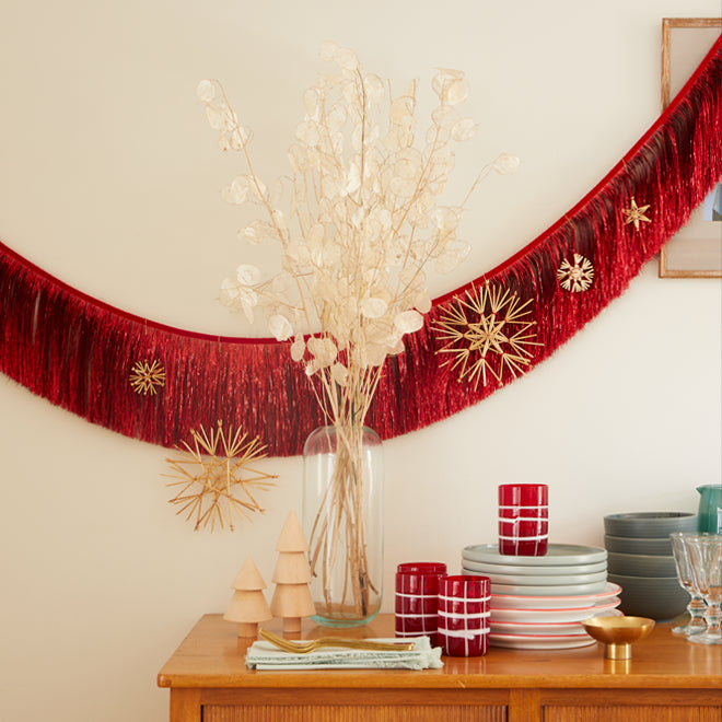 Stacks of blue and red dinnerware and glassware on a wood sideboard. In the center a vase of dried florals. Above hangs a swag of red metallic fringed garland decorated with straw ornaments.